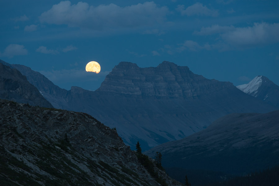 The moon rises above mountains and clouds on a warm summer evening.
