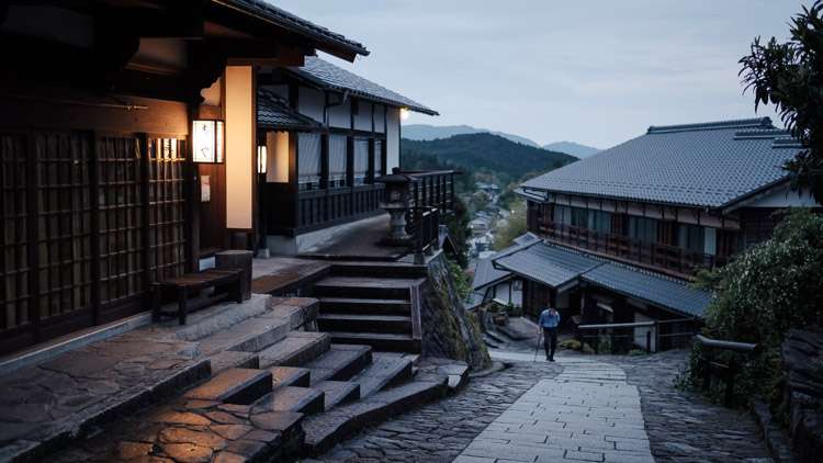 A person with a walking stick climbs the stone street of Magome at dusk, lantern light glowing on old wooden inns with mountains beyond