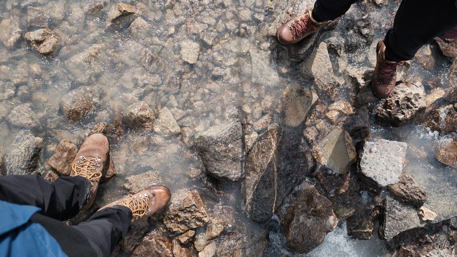 Looking down at two hikers' leather boots crossing the clear, rocky shallows of the Brazeau River