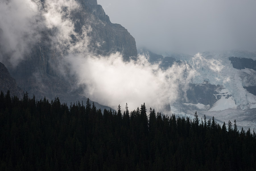 Clouds drifting across cliff faces and the blue ice of the Columbia Icefield, dark treeline silhouetted below