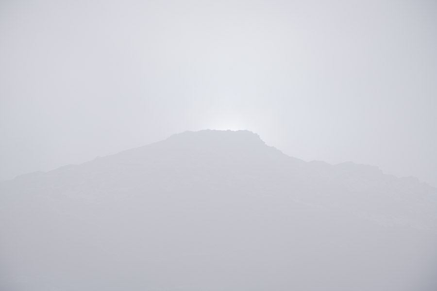 A mountain ridge reduced to a faint grey silhouette in dense fog, the sun a pale glow just behind its summit
