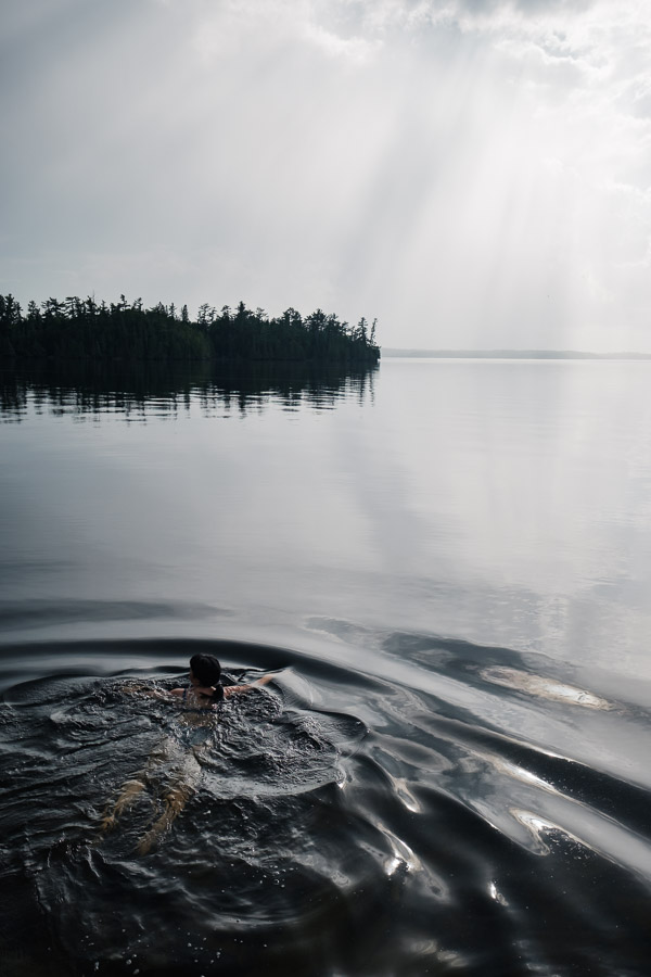 A swimmer strokes out into a glassy lake toward a pine-covered point, sunrays breaking through haze