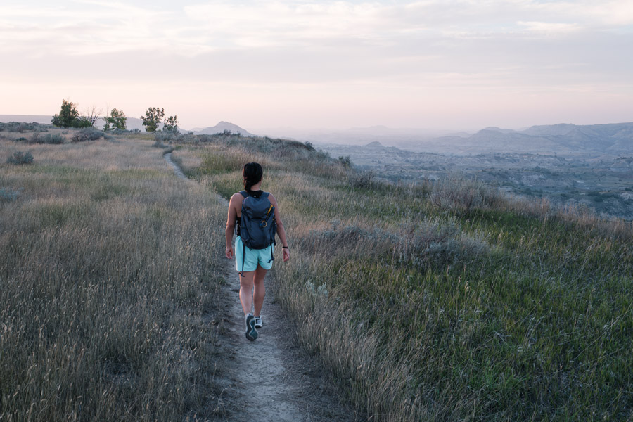 A hiker with a small backpack follows a grassy trail along the rim above the hazy pastel buttes of Painted Canyon