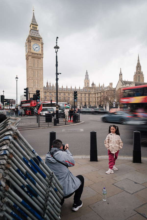 A crouching photographer takes a young girl's portrait on a corner by Big Ben while buses and cars streak past