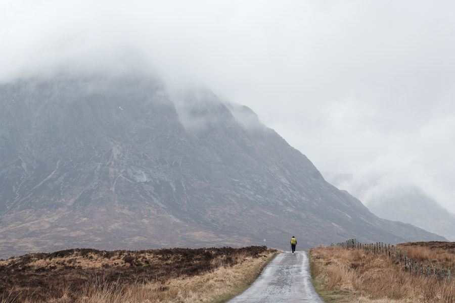 A lone hiker in a yellow pack cover walks a wet road dwarfed by the mist-shrouded face of Buachaille Etive Mor