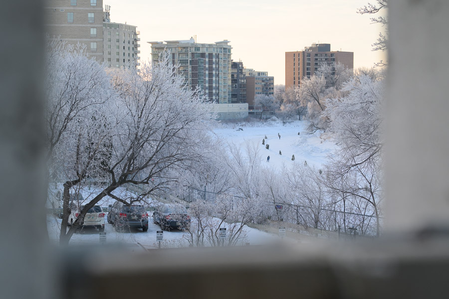Framed by a concrete overlook, skaters glide on the distant river between frost-covered trees and morning-lit apartment blocks